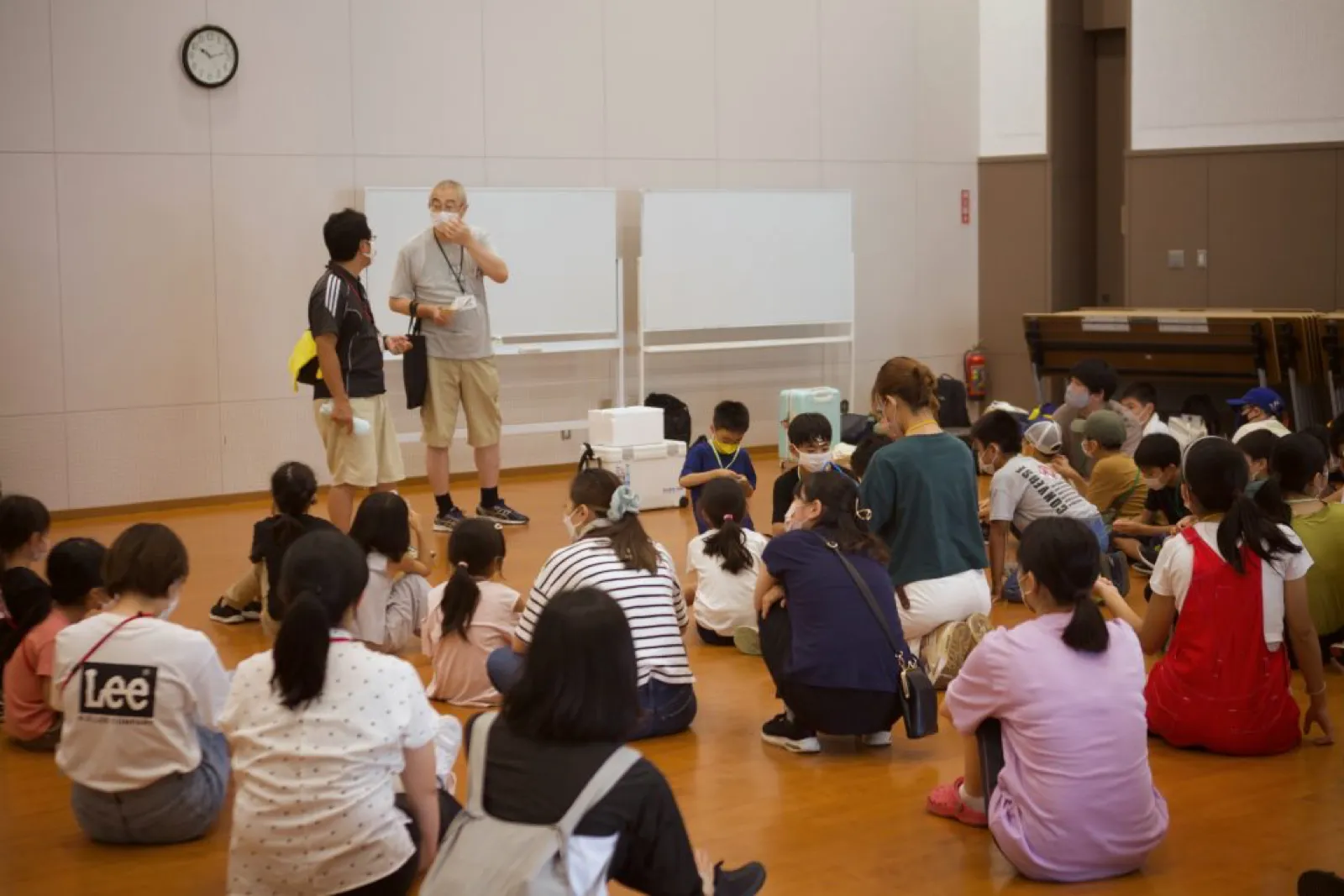 退村式で、体育館の床に座った子どもたちとスタッフが、前に立つ二人の話を聞いている。