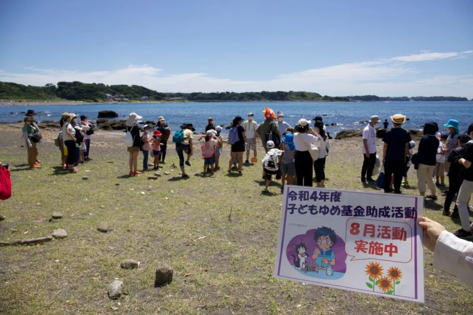 磯遊びの前に、海辺の草地で子どもとスタッフが輪になって集まっている。奥に入り江が広がる。