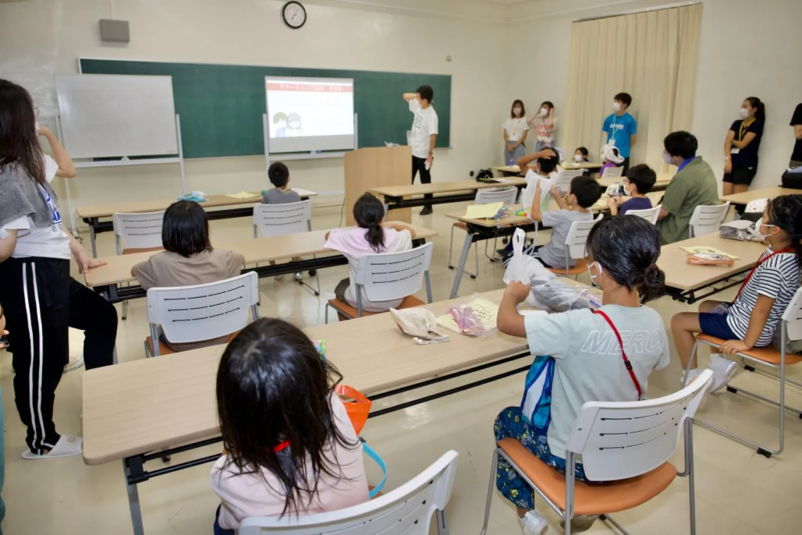 夕食後の学習会で、教室の机に座った子どもたちが、スクリーンの資料を見ながら手元の道具を扱っている。