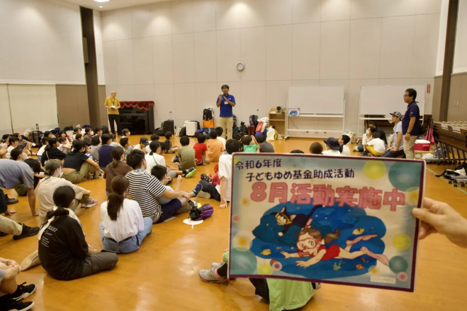 退村式で、体育館の床に座った子どもたちが、前に立つスタッフの話を聞いている。