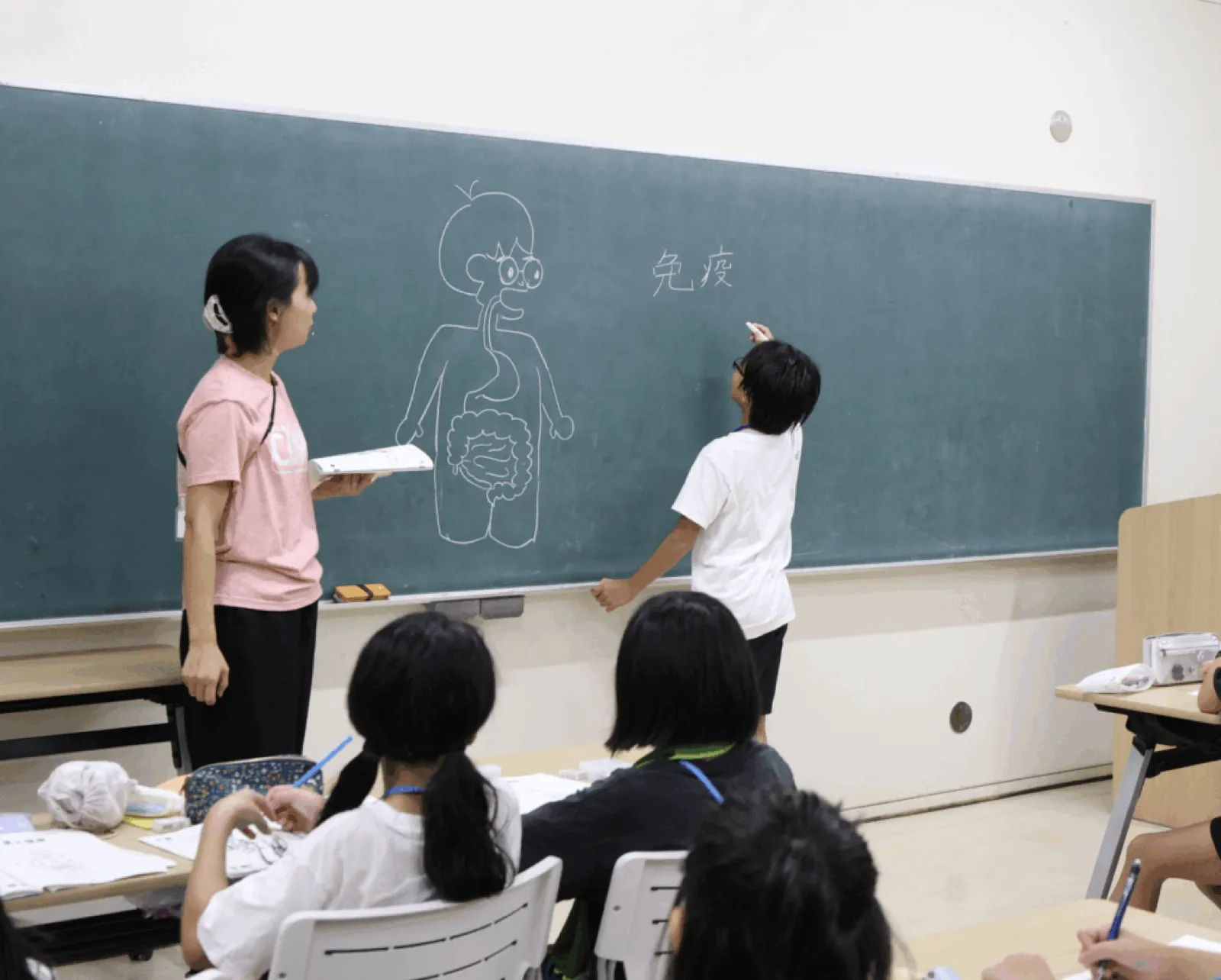 食物アレルギーの学習会で、黒板の体の絵と「免疫」の文字の前に、子どもが出て書き込み、スタッフが見守っている。